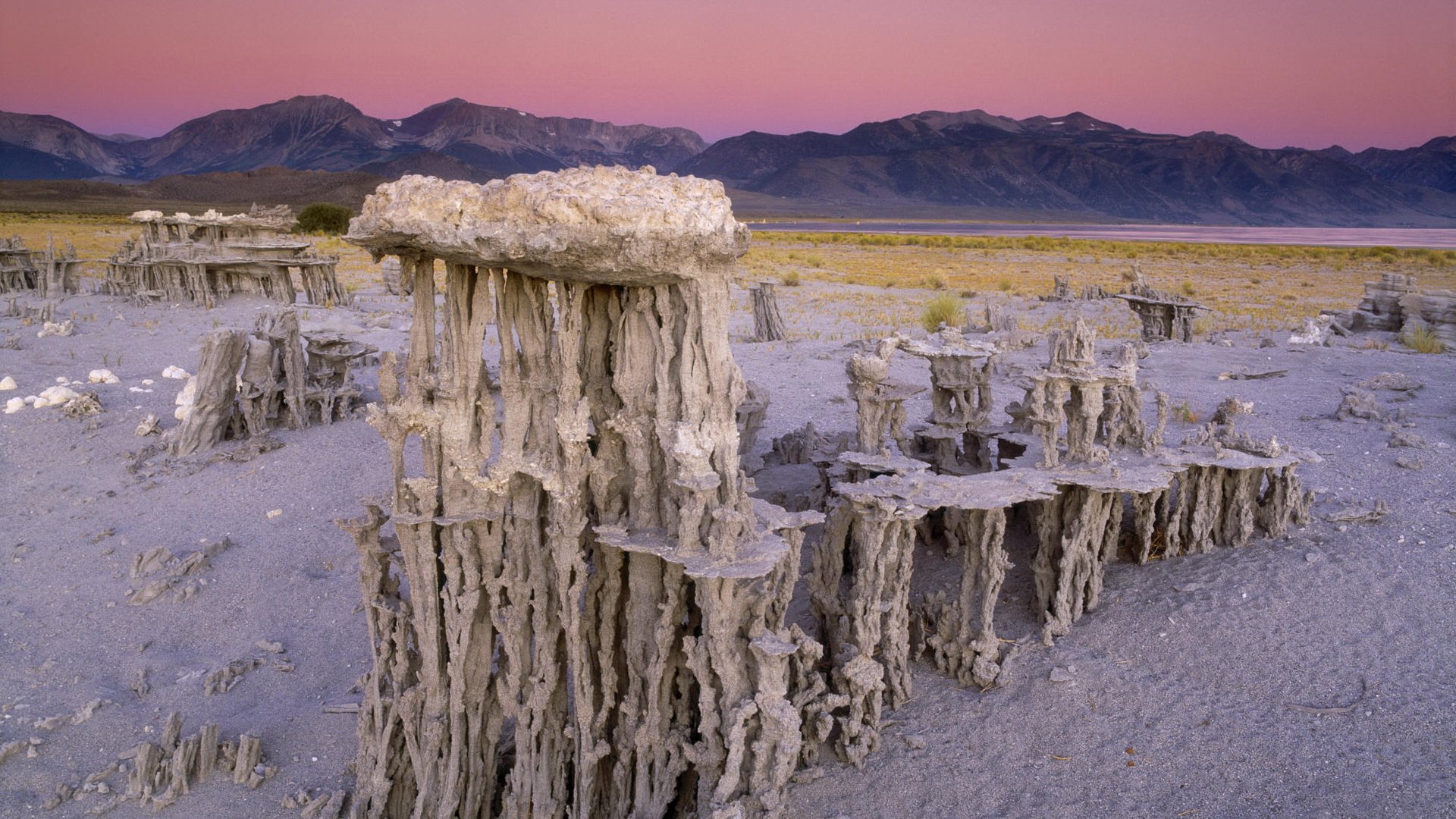 HD PC desktop wallpaper showcasing unique desert rock formations with a pastel sunset sky and distant mountain range in the background.
