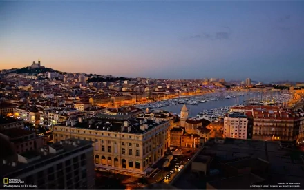 HD desktop wallpaper showcasing the illuminated man-made skyline of Marseille at dusk, featuring the harbor and city buildings under a clear evening sky.