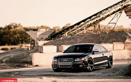 HD PC desktop wallpaper featuring a sleek black Audi S5 parked in an industrial setting with gravel and machinery in the background.