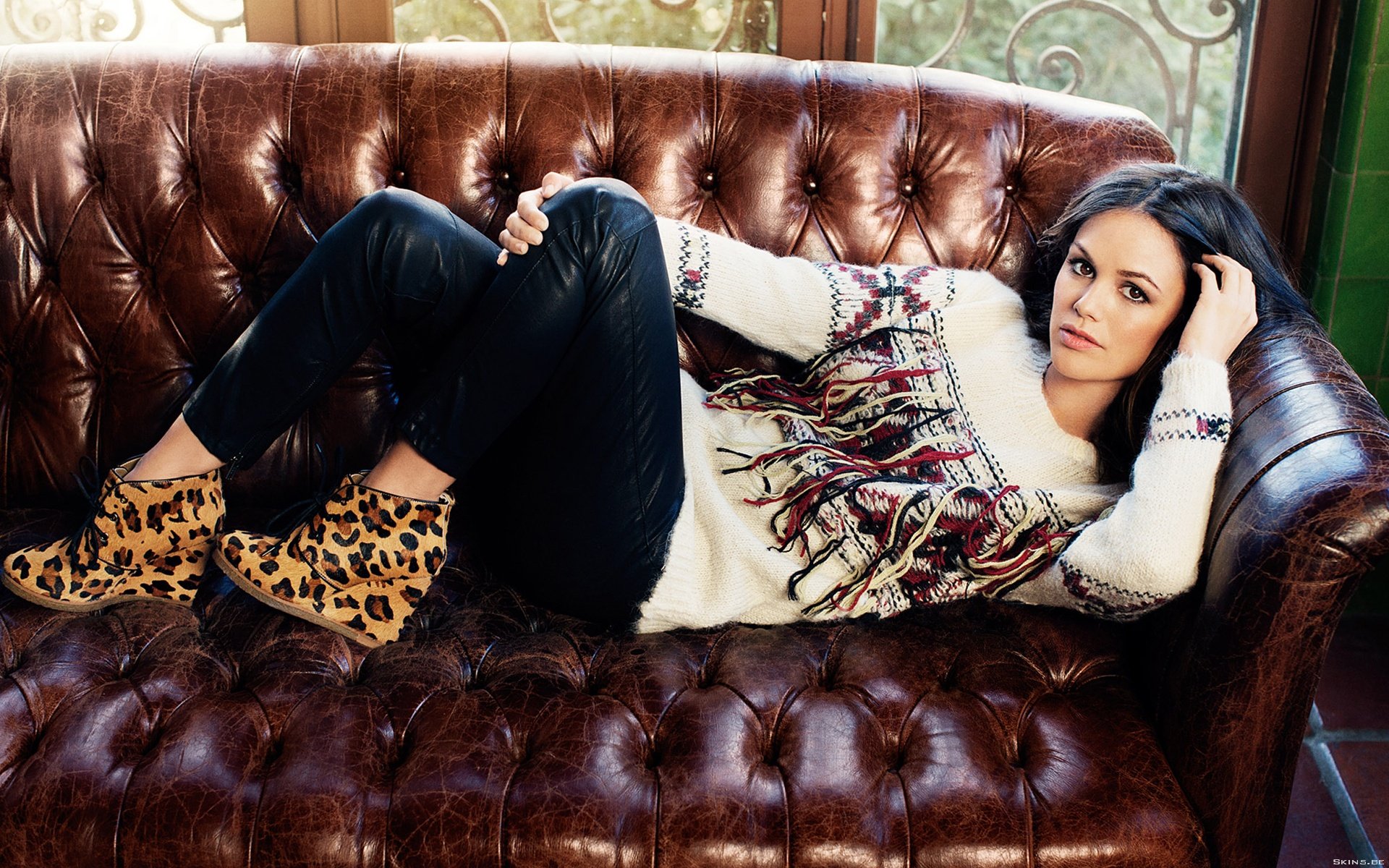 HD desktop wallpaper featuring celebrity Rachel Bilson reclining on a tufted leather sofa, dressed in a patterned sweater, black pants, and leopard print shoes.