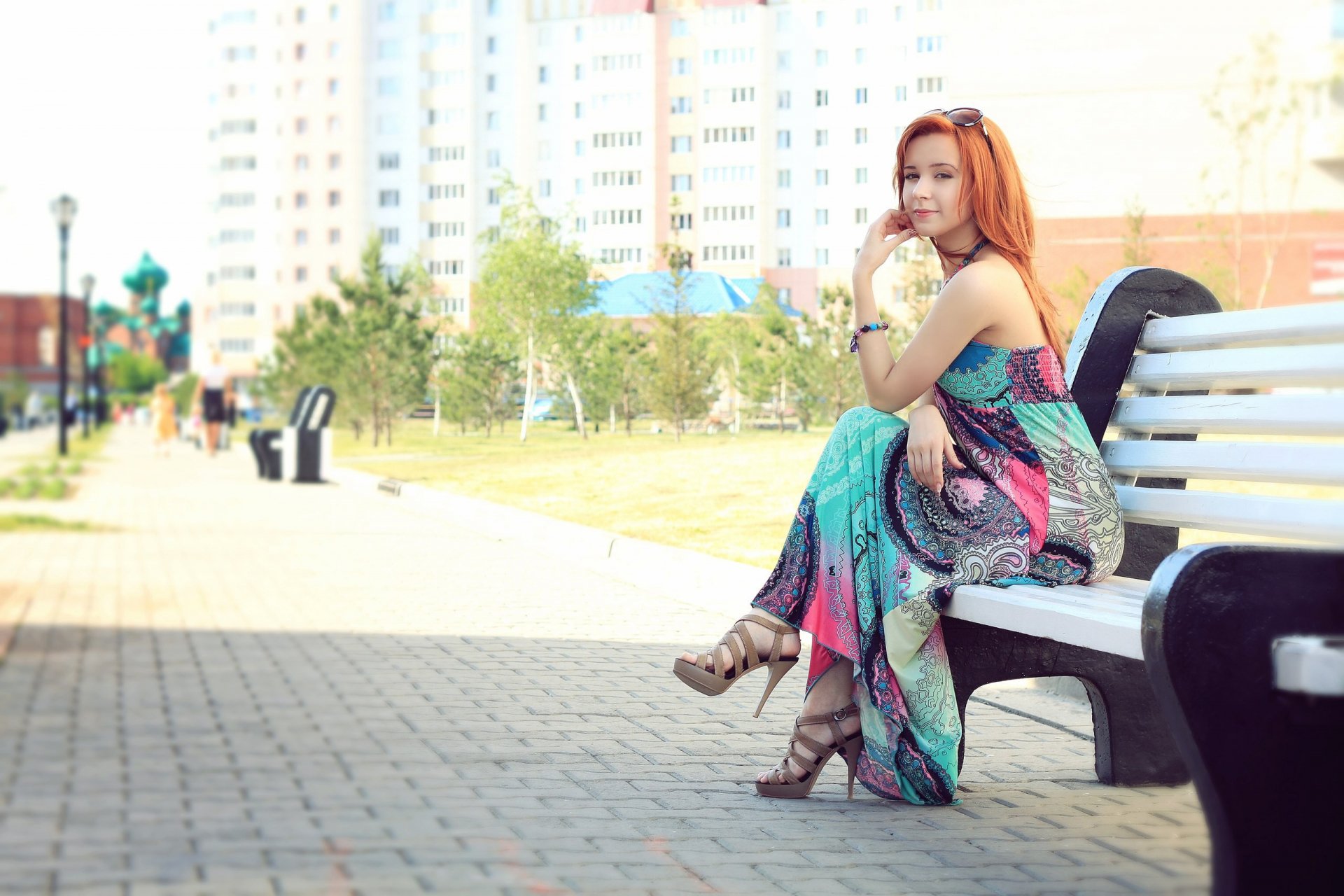 A woman model with red hair sits gracefully on a white bench in an urban park. She wears a colorful, flowing dress, creating an elegant and serene scene. This HD wallpaper and background exudes charm and sophistication.