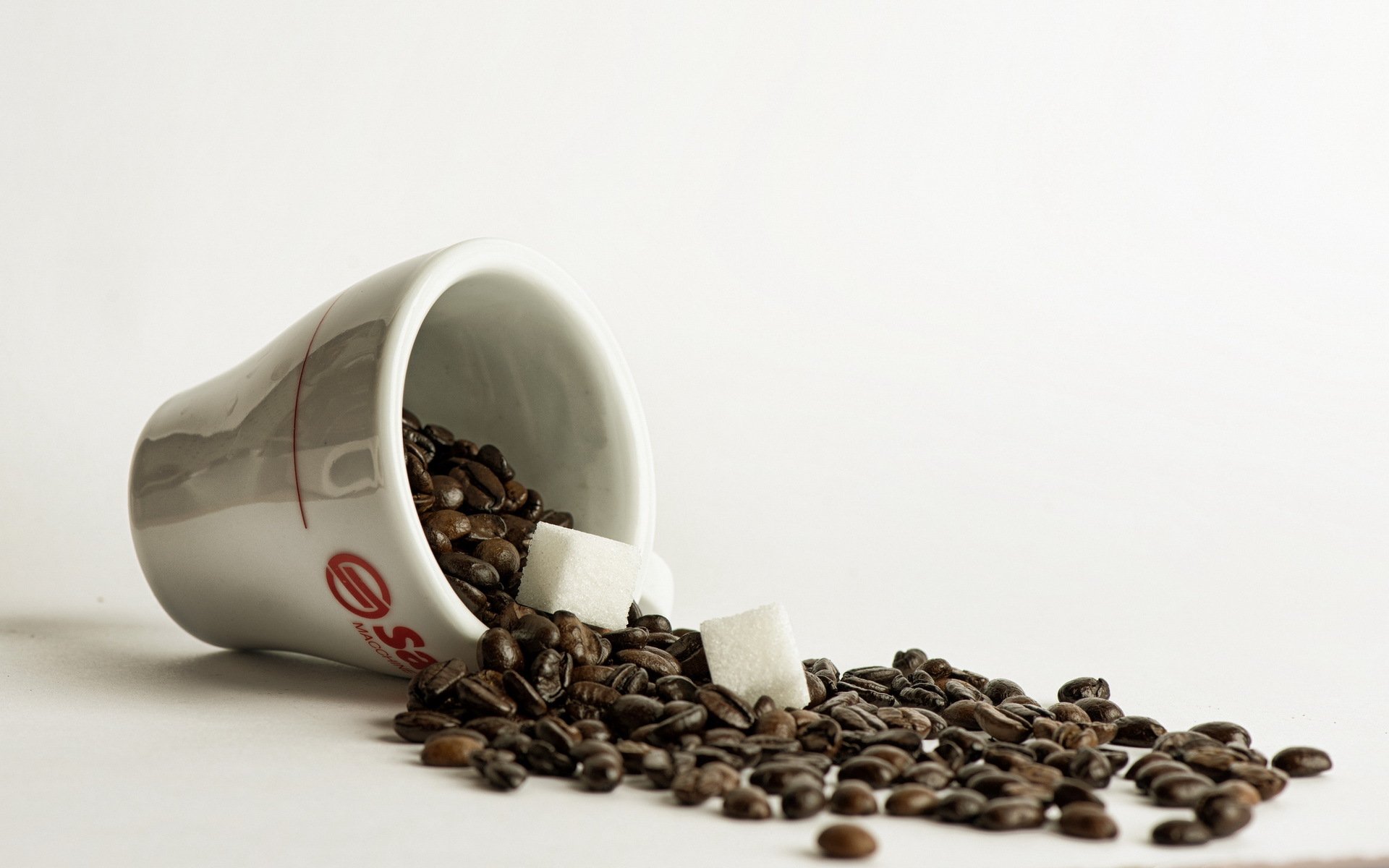 HD PC desktop wallpaper showing a white cup spilled with coffee beans and two sugar cubes on a clean white background.