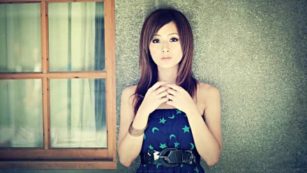 A woman named Mikako Zhang Kaijie poses in front of a window, wearing a star-patterned dress and an elegant belt, against a textured wall, creating a serene and stylish image.