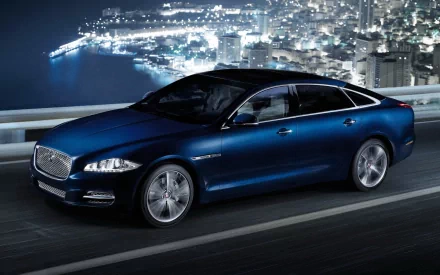 HD PC desktop wallpaper: sleek blue Jaguar XJ sedan cruising on a coastal highway at night with an illuminated city skyline in the background.
