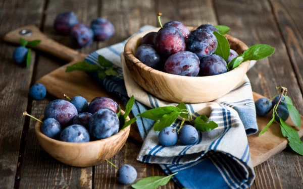 HD PC desktop wallpaper showing ripe plums in wooden bowls on a rustic table, with green leaves and a blue-striped cloth for a vibrant food background.