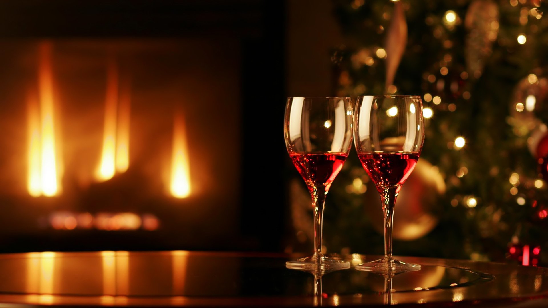 Two glasses of red wine sit on a table, reflecting warm light from a fireplace, with a festive tree adorned in the background. A cozy and inviting scene for a desktop wallpaper.