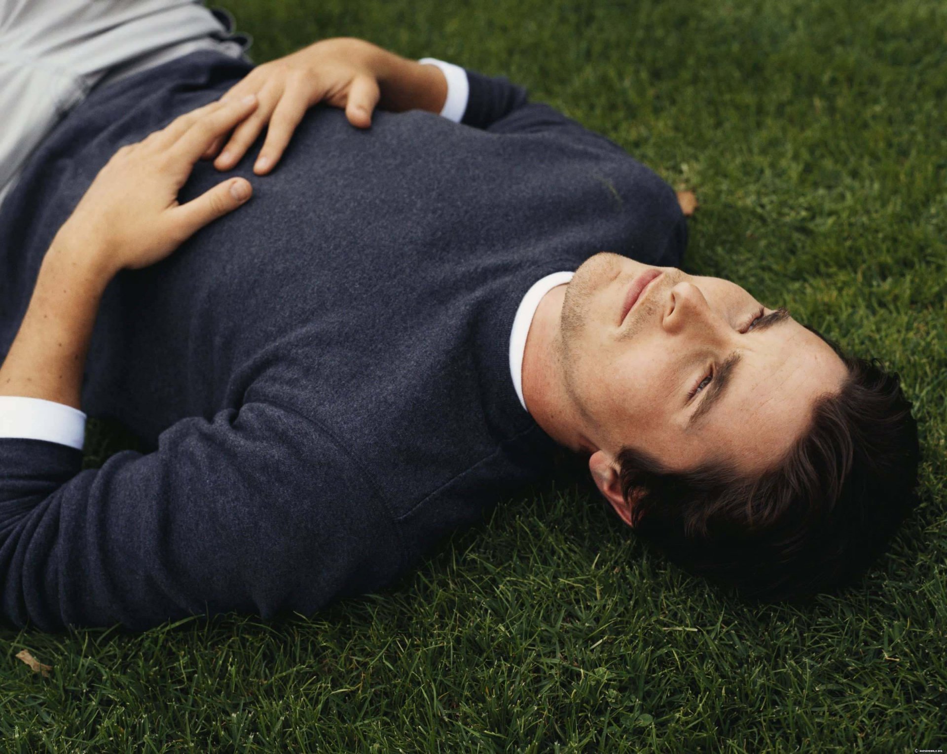 HD PC desktop wallpaper featuring celebrity Christian Bale lying on grass with eyes closed, dressed in a dark sweater over a white shirt.