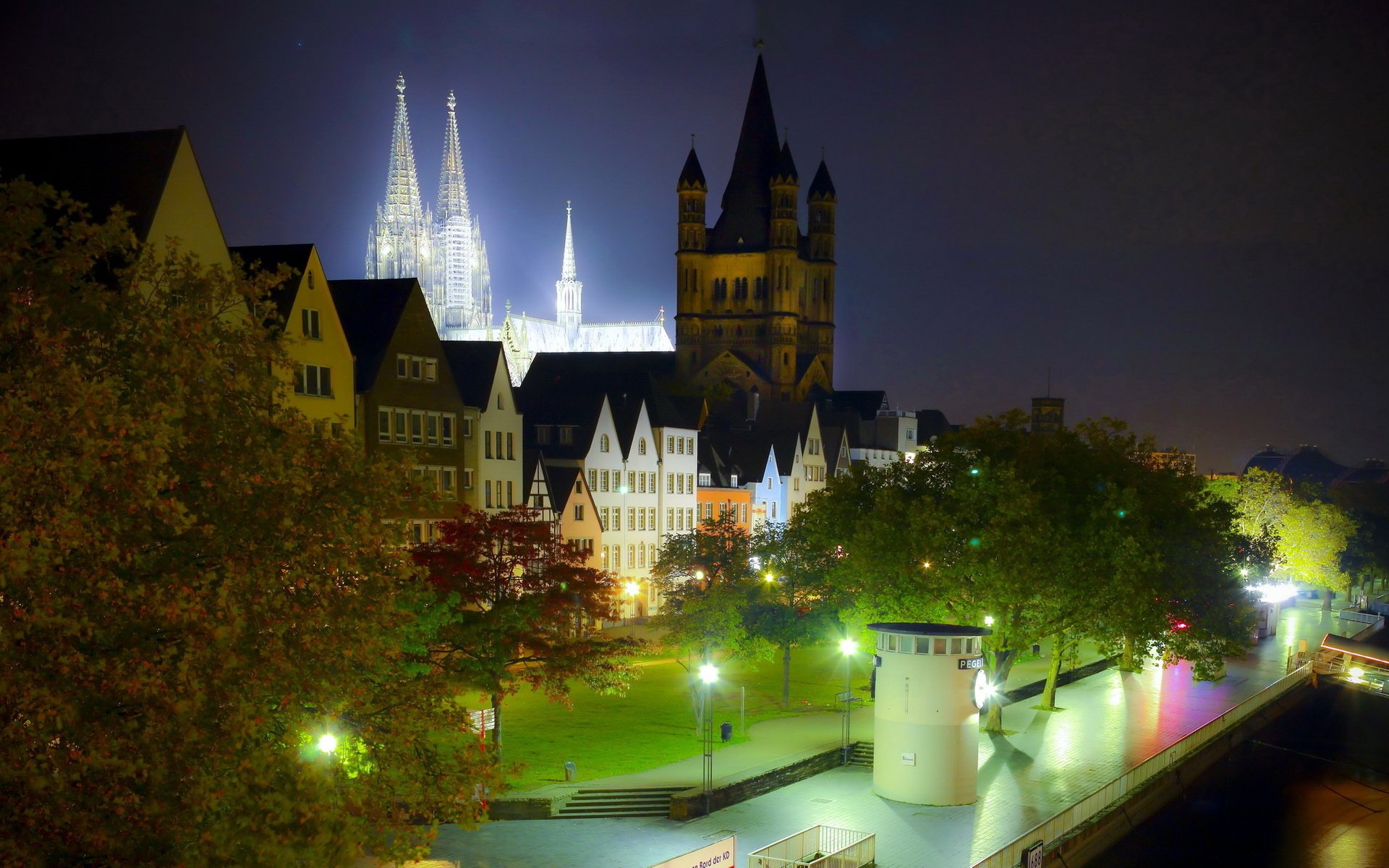 HD PC desktop wallpaper: nighttime view of man-made Cochem Castle and illuminated historic riverside buildings, tree-lined promenade and shimmering water reflections.