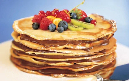 HD desktop wallpaper of a tall stack of crêpes layered with chocolate spread, topped with strawberries, blueberries and kiwi against a soft blue background.