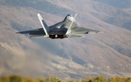 A Lockheed Martin F-22 Raptor takes flight over mountainous terrain, showcasing its sleek design and advanced military capabilities in this HD desktop wallpaper.