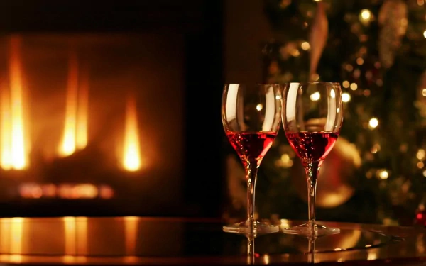 Two glasses of red wine sit on a table, reflecting warm light from a fireplace, with a festive tree adorned in the background. A cozy and inviting scene for a desktop wallpaper.