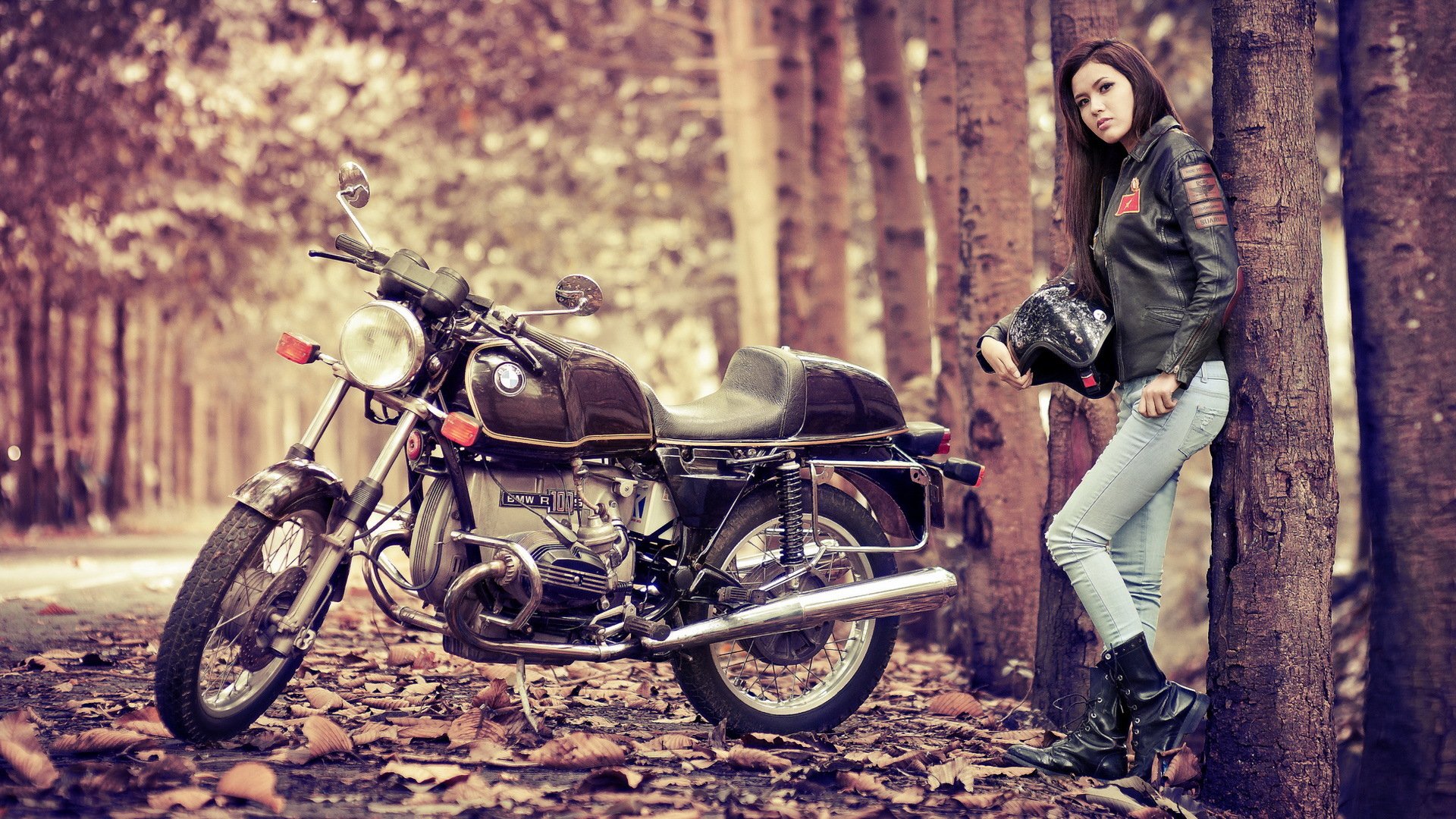 A woman in a leather jacket stands by a vintage motorcycle in an autumn forest, creating a stylish and moody HD desktop wallpaper and background.
