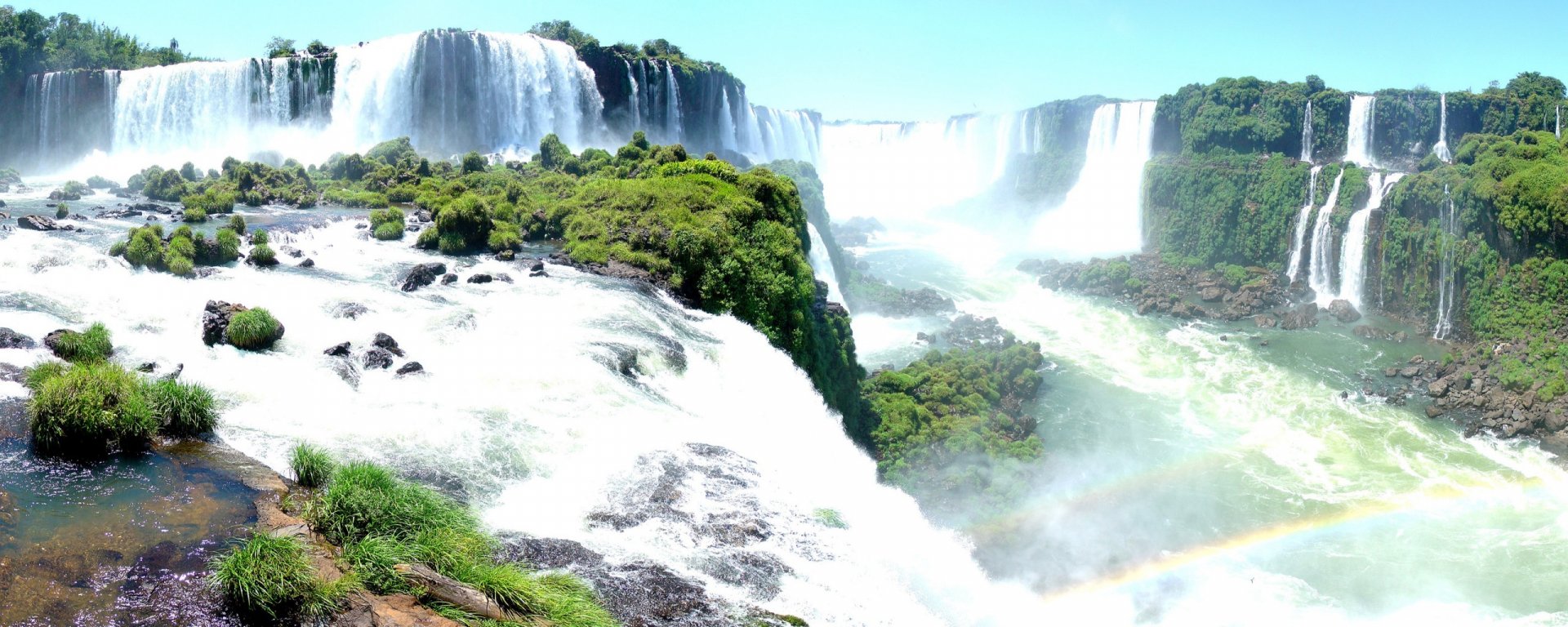 HD desktop wallpaper showcasing the powerful Iguazu Falls surrounded by lush greenery and mist under a clear sky, capturing the beauty of nature.