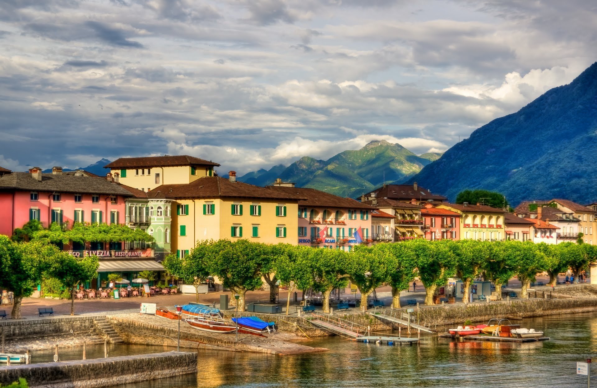 HD PC desktop wallpaper/background: vibrant man-made village along a lakeshore with colorful buildings, boats, a tree-lined promenade and mountains beneath a cloudy sky.