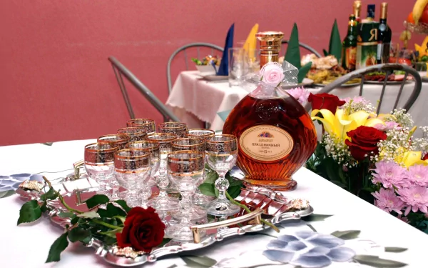 A beautifully arranged table features a bottle of liquor surrounded by decorative glasses, complemented by vibrant flowers and a soft pink backdrop, creating an inviting and festive atmosphere.