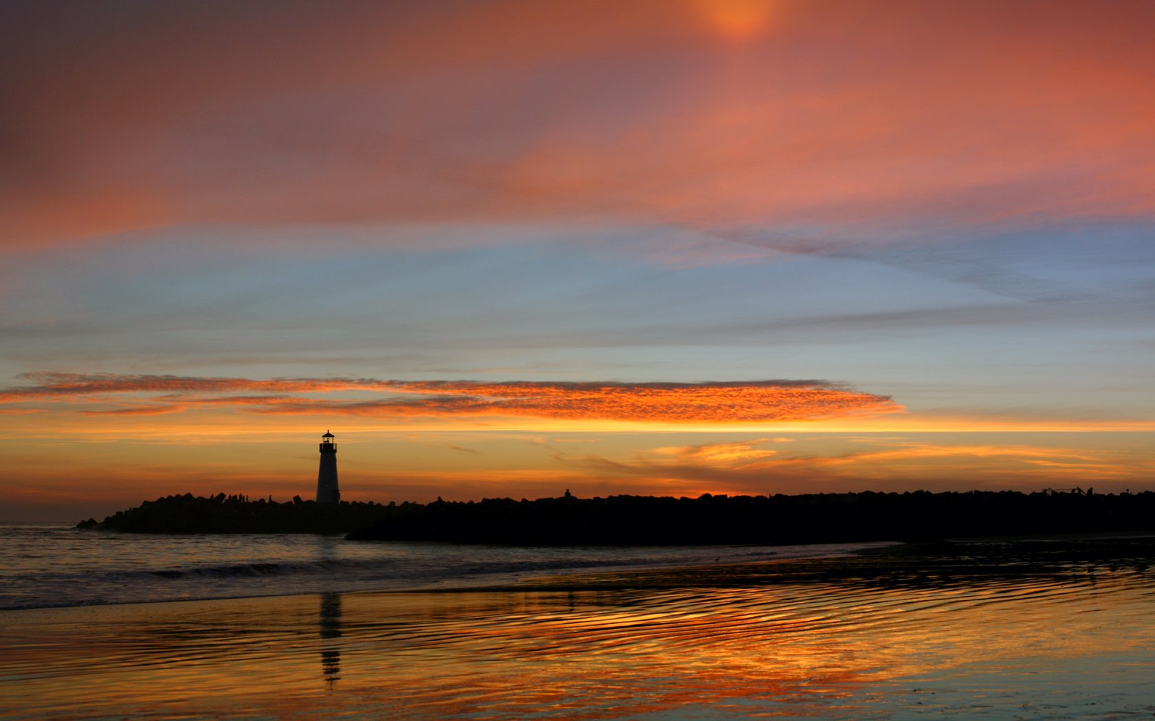 HD PC desktop wallpaper showing a man-made lighthouse silhouette against a vivid sunset, warm sky reflected across calm water.
