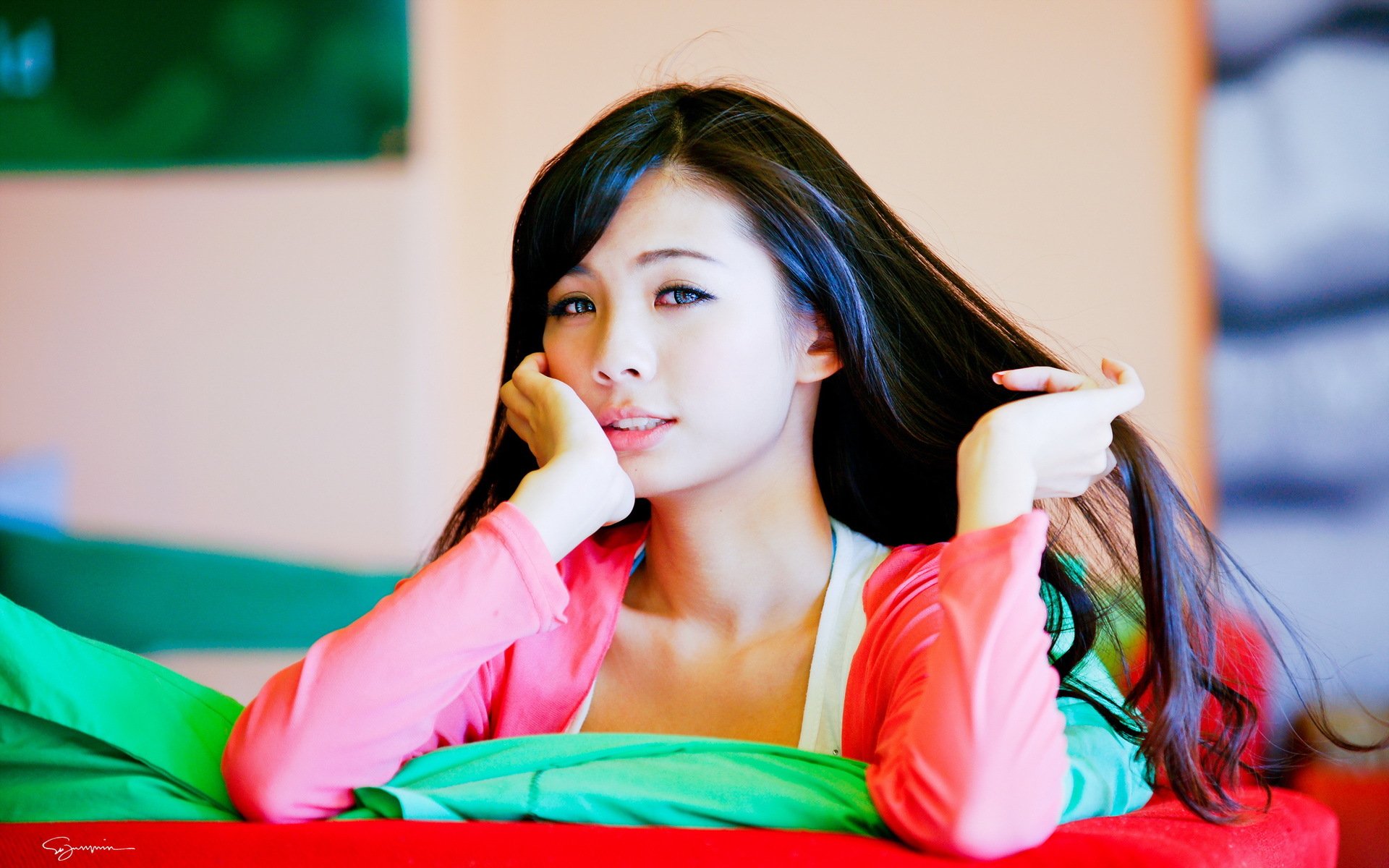 A serene Asian woman poses with her long hair, resting her chin on her hand, set against a colorful background. This HD image makes for an engaging desktop wallpaper.