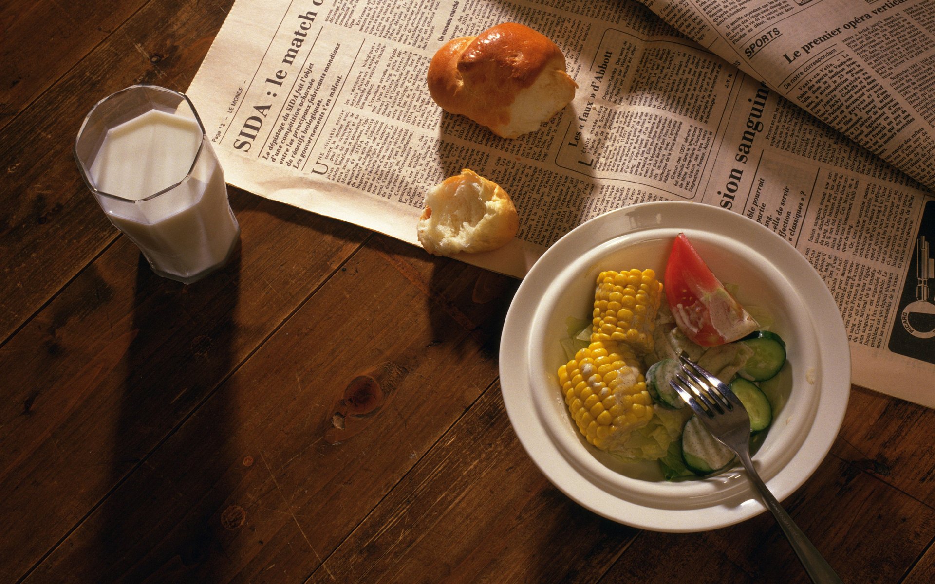 HD PC desktop wallpaper featuring a meal with corn on the cob, sliced vegetables, bread rolls, and a glass of milk on a wooden table with a newspaper.