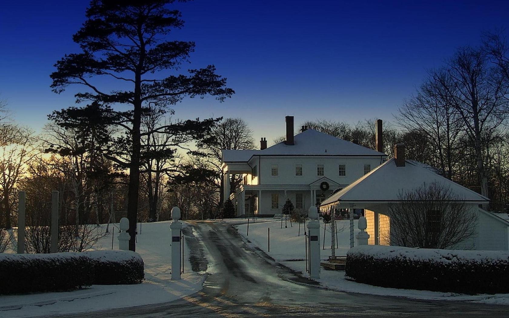 HD PC desktop wallpaper: snow-covered man-made mansion at dusk, lit driveway flanked by tall trees.