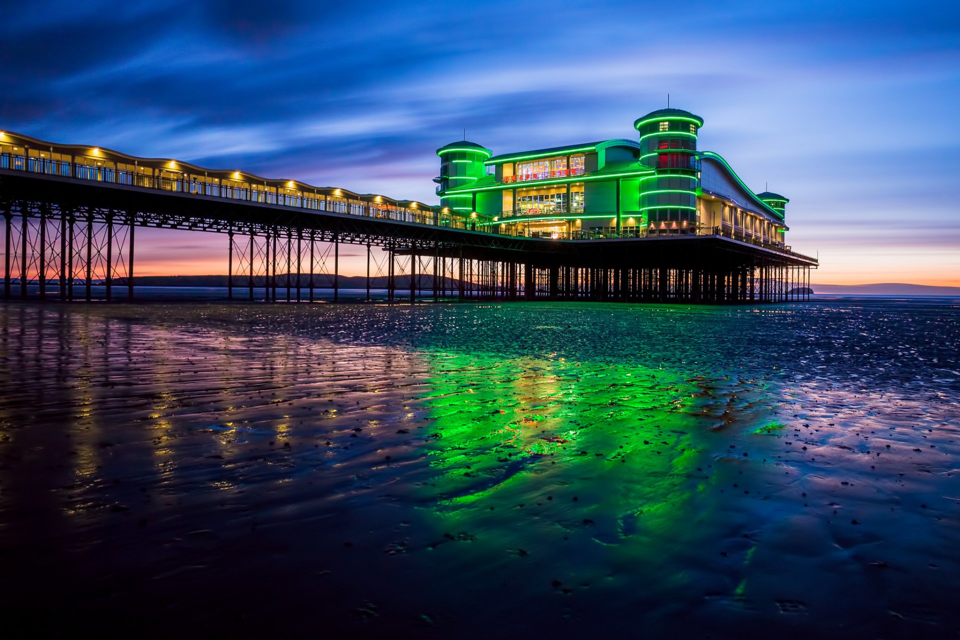 HD desktop wallpaper of a brightly lit man-made pier extending over calm water at twilight, with vibrant green reflections shimmering on the wet sand below.