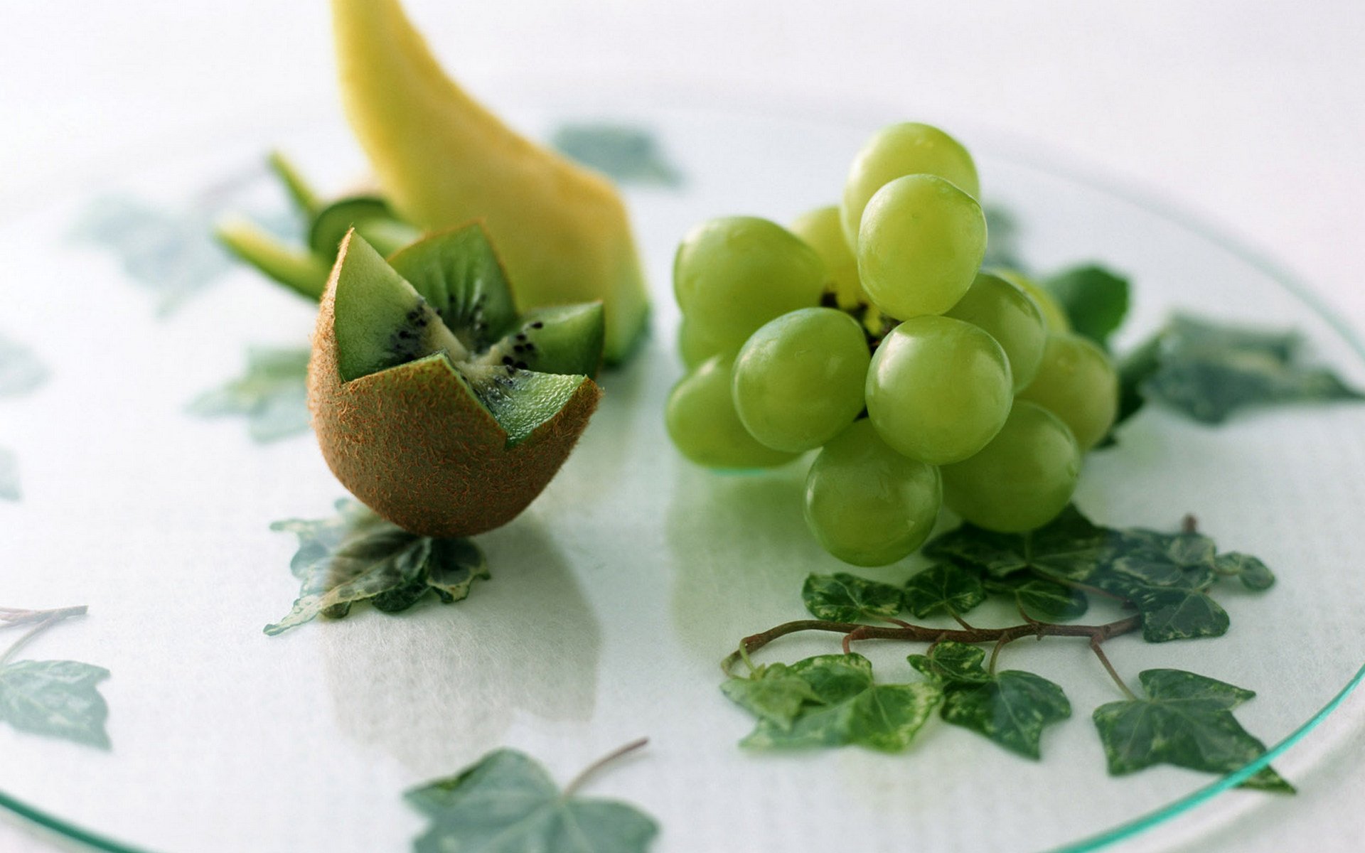 HD PC desktop wallpaper background of food/fruit: close-up of a halved kiwi, a bunch of green grapes and a curved banana on a glass plate with an ivy-patterned surface.