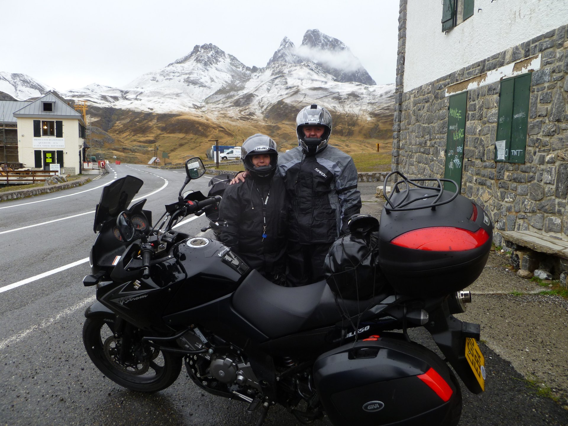 Two people in motorcycle gear stand beside a black Suzuki V-Strom on a mountain road, with snow-capped peaks and a village in the background, captured in 4K Ultra HD.