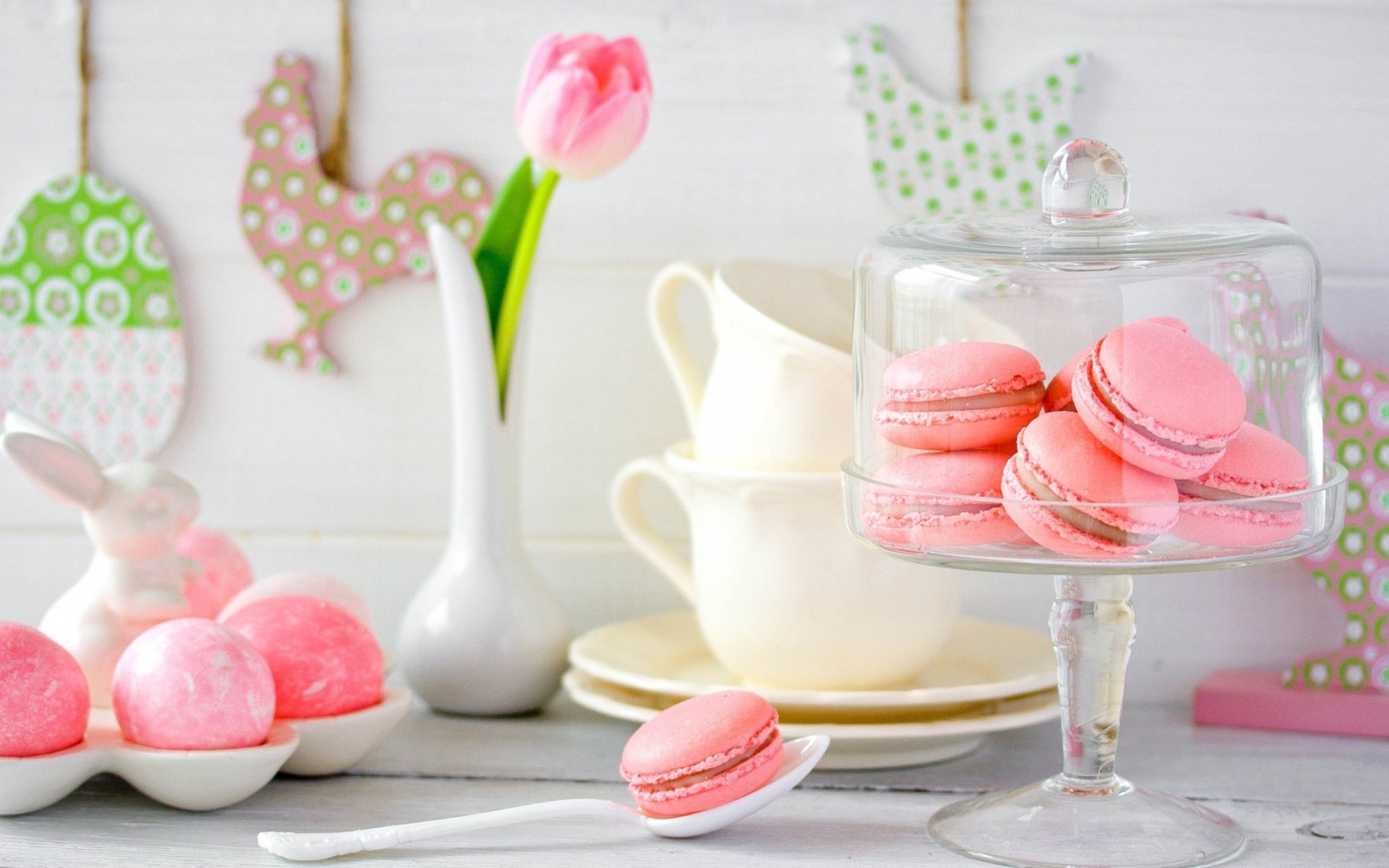 A vibrant display of pink macarons in a glass dome, complemented by elegant tableware and a tulip vase, creating a charming food-themed wallpaper for a desktop background.