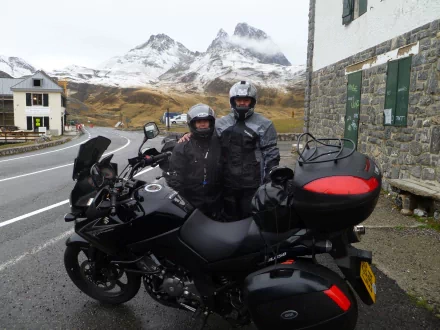 Two people in motorcycle gear stand beside a black Suzuki V-Strom on a mountain road, with snow-capped peaks and a village in the background, captured in 4K Ultra HD.
