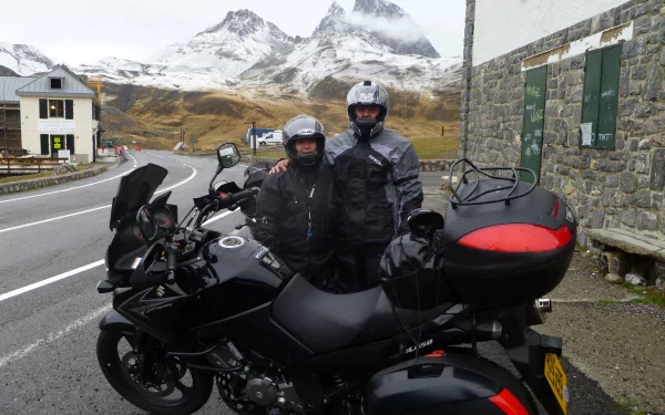 Two people in motorcycle gear stand beside a black Suzuki V-Strom on a mountain road, with snow-capped peaks and a village in the background, captured in 4K Ultra HD.