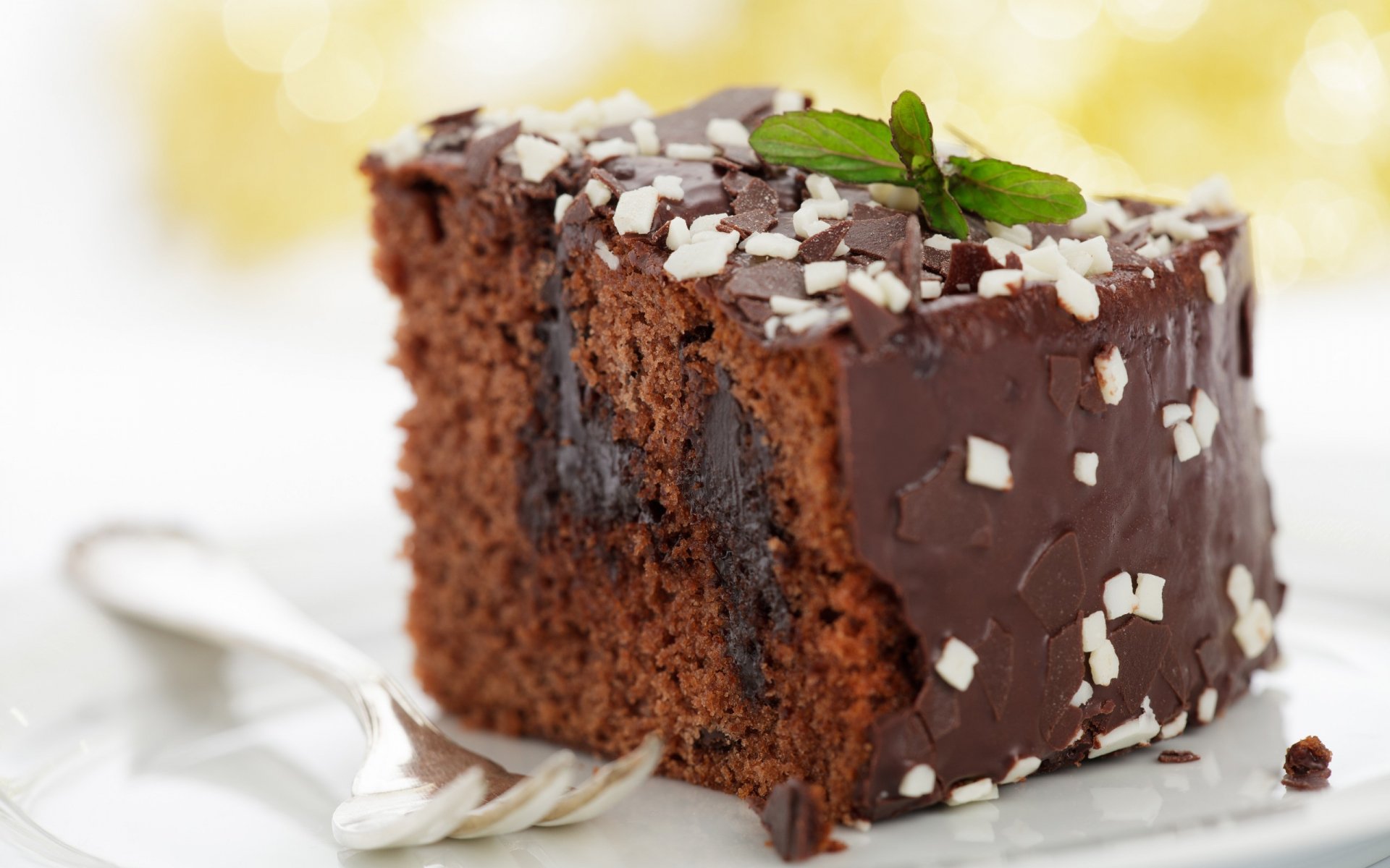 HD desktop wallpaper featuring a rich chocolate cake slice topped with white chocolate shavings and a fresh mint leaf on a blurred light background.
