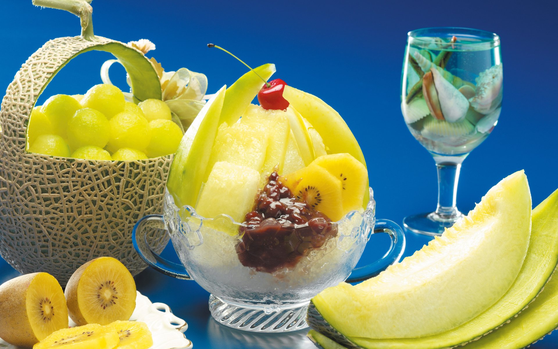 HD PC desktop wallpaper featuring a vibrant arrangement of fresh fruit including melon, grapes, kiwi, and a glass with fruit pieces against a bright blue background.