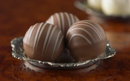 A close-up of three exquisite chocolate truffles with white drizzle, elegantly presented on a silver plate, serves as a deliciously inviting HD PC desktop wallpaper and background.