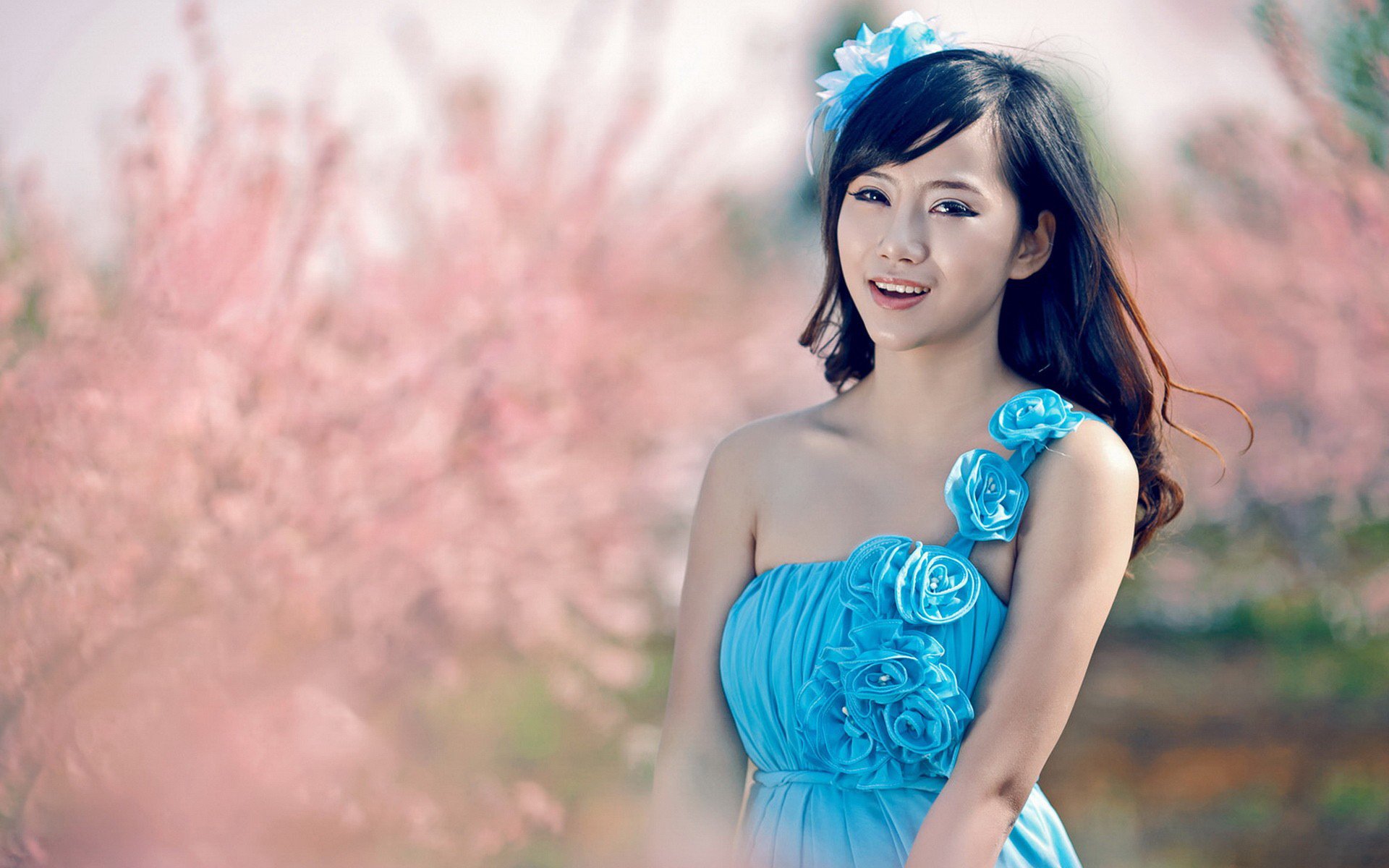 A smiling Asian woman in a blue dress stands among soft pink blossoms, captured in a vibrant HD desktop wallpaper.