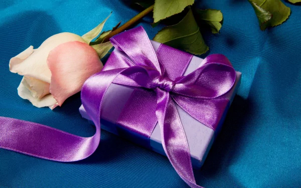 HD PC desktop wallpaper featuring a purple gift box with a satin ribbon next to a pale pink rose on a vibrant blue fabric background.