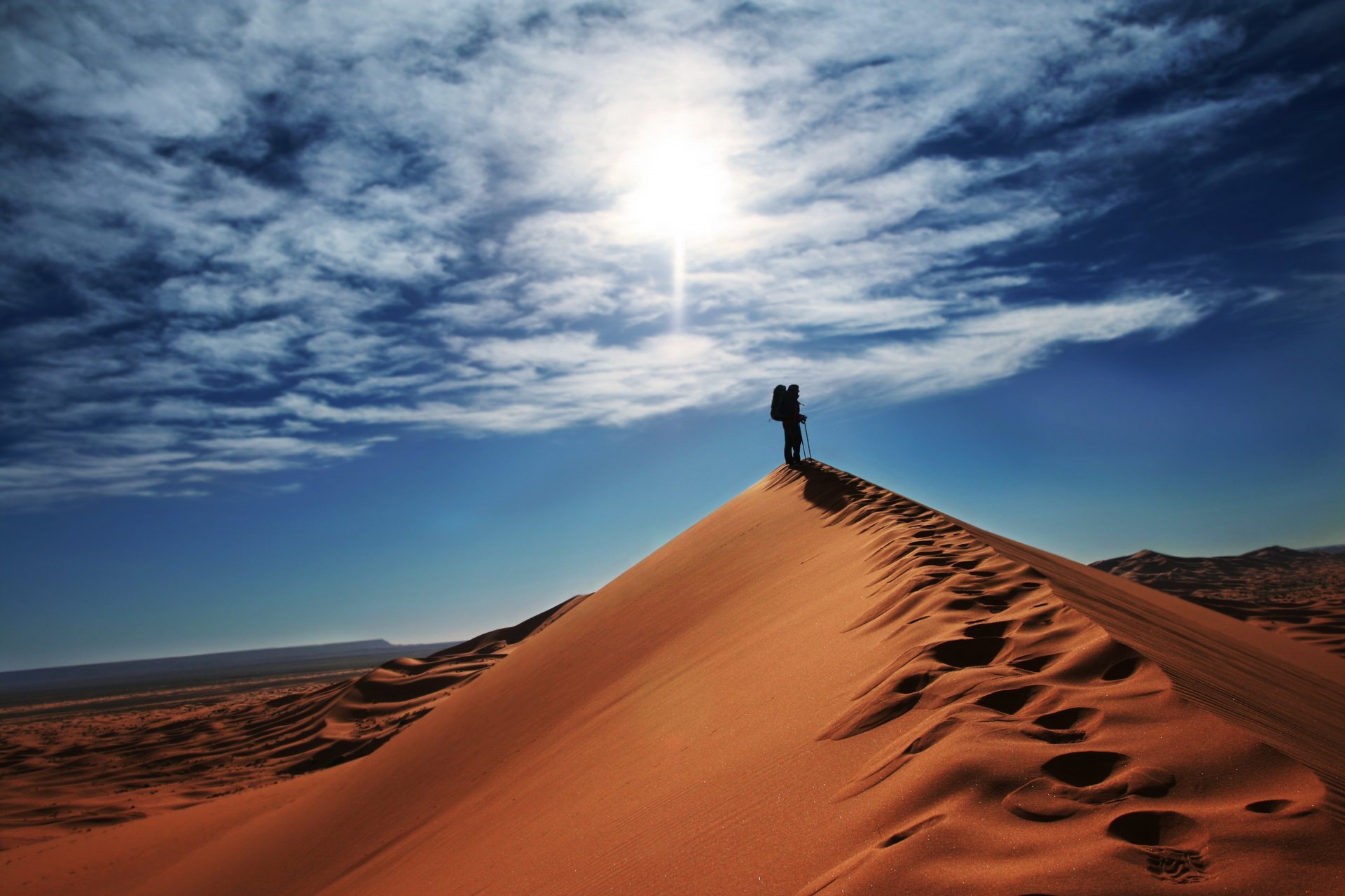 2K Quad HD PC desktop wallpaper: lone figure standing on a desert dune in a dramatic nature scene beneath a bright sun and sweeping clouds, footprints trailing down the ridge.