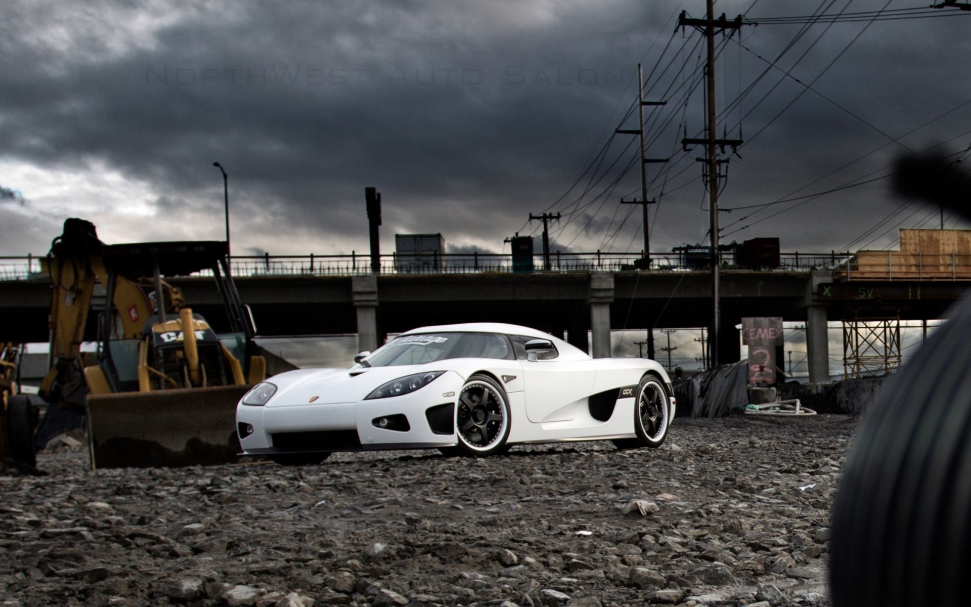 HD desktop wallpaper featuring a white Koenigsegg CCX sports car parked on a gravel surface with industrial structures and a cloudy sky in the background.
