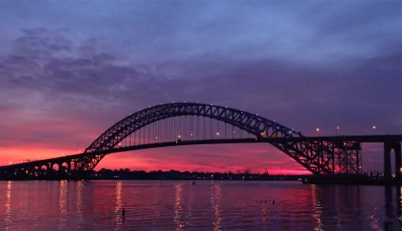 4K Ultra HD wallpaper showing the man-made Bayonne Bridge silhouetted against a vibrant sunset sky over calm water.