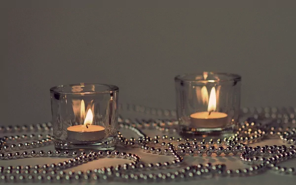 HD desktop wallpaper featuring a photography shot of two lit candles in glass holders surrounded by silver bead chains on a neutral background.