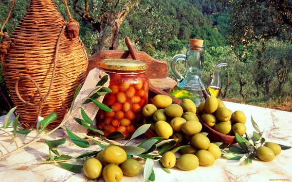 HD desktop wallpaper showing a rustic outdoor table with a jar of preserved olives, fresh olives, a glass bottle of olive oil, and a woven basket in an olive grove setting.