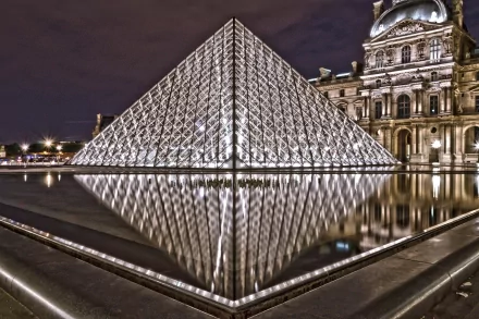 Night view of the illuminated glass pyramid at The Louvre in Paris, France, reflecting on the water, captured in HD as a striking man-made architectural scene.
