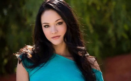 A confident woman with long dark hair and a teal top stands against a natural green background in this HD desktop wallpaper.