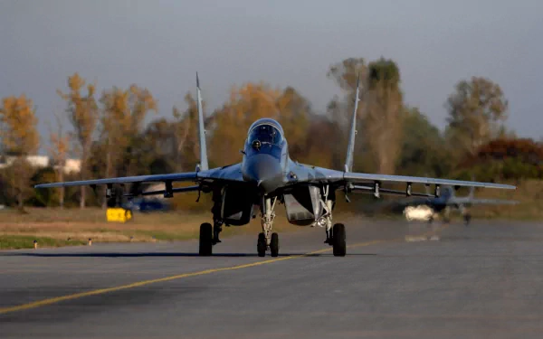 A Mikoyan MiG-29 jet fighter taxies on a runway, showcasing its military design against a backdrop of autumn foliage. This high-definition image serves as a striking desktop wallpaper.