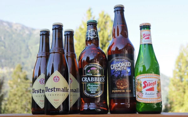 HD desktop wallpaper featuring a selection of beer bottles, including craft and seasonal brews, set against a blurred outdoor background with trees and mountains.