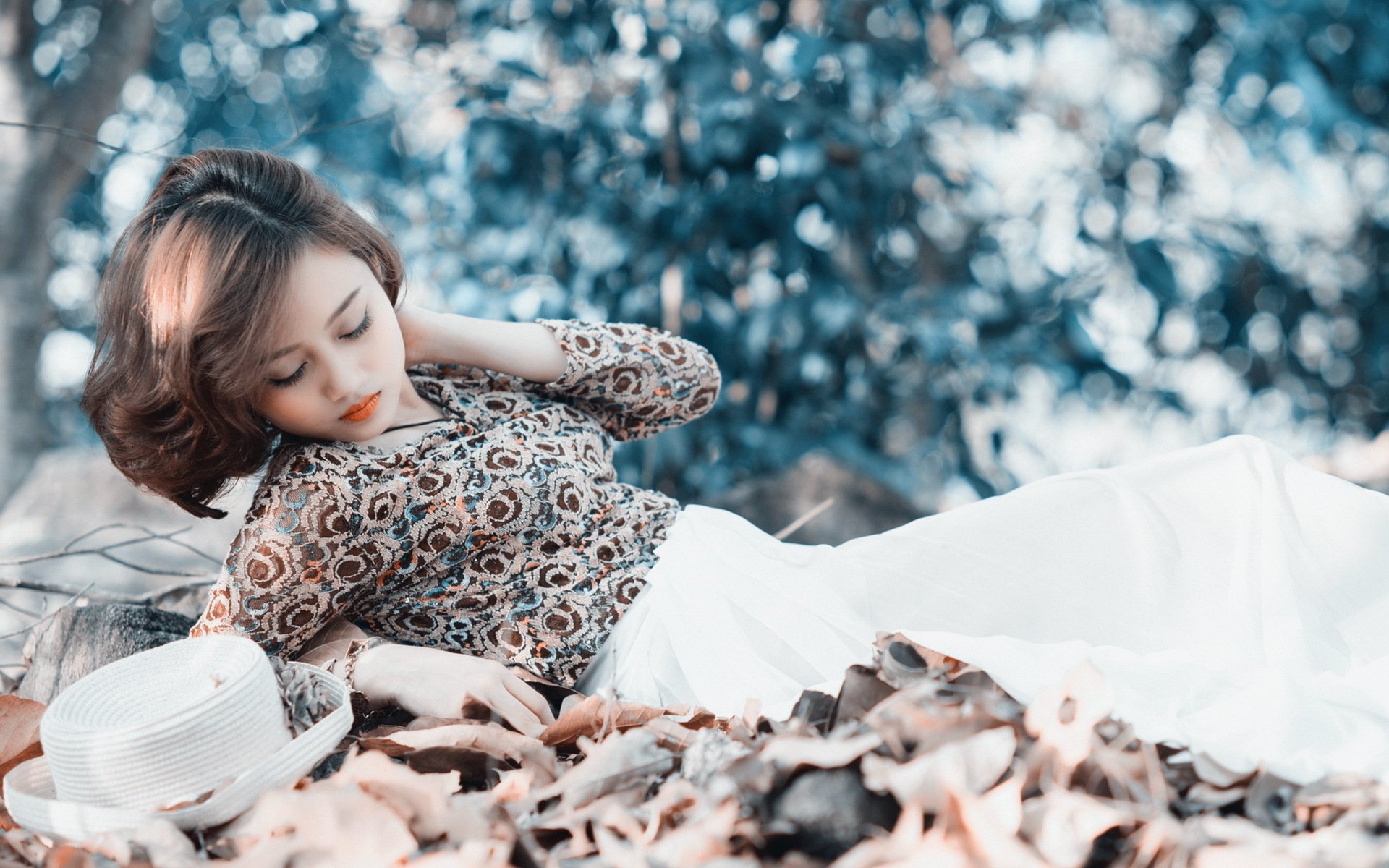 Asian woman in a patterned blouse and white pants lies on fallen leaves in a serene, blue-toned woodland. A white hat is placed beside her. This HD desktop wallpaper captures a peaceful outdoor moment.