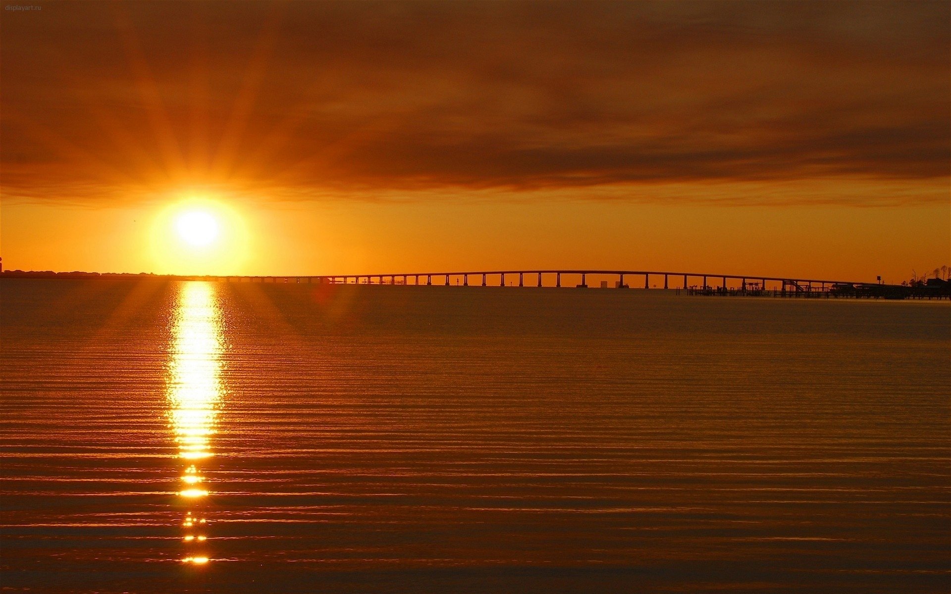 HD PC desktop wallpaper/background photography: golden sunset over calm water, sun's bright reflection leading to a distant low bridge silhouette under a warm, glowing sky.