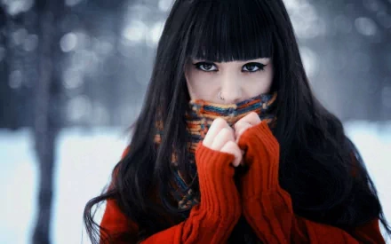 HD desktop wallpaper featuring a pretty woman with long dark hair, wearing a red sweater and colorful scarf against a snowy, blurred background.