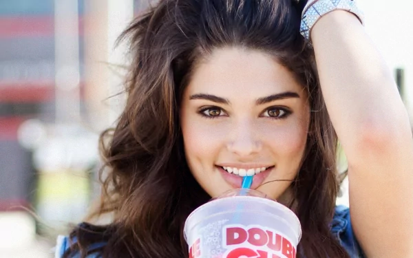 Bright 2K Quad HD PC desktop wallpaper of a smiling young woman sipping from a drink, tousled hair and hand raised, with a softly blurred outdoor background.