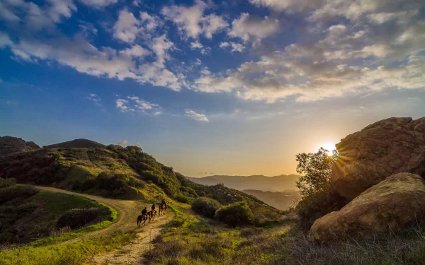 HD desktop wallpaper photography capturing a sunbeam breaking through clouds over rolling hills with a group of hikers on a winding trail.
