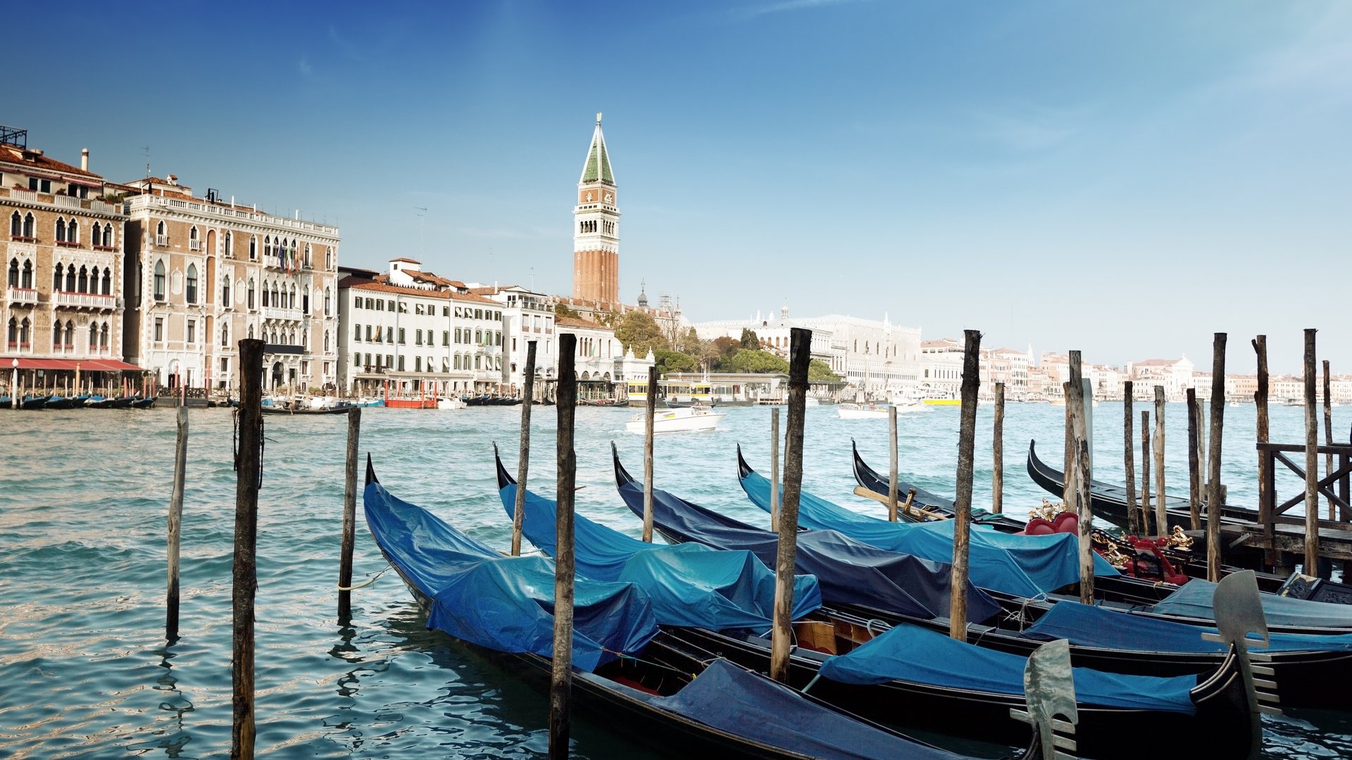HD PC desktop wallpaper of man-made Venetian waterfront: gondolas moored to wooden posts, historic buildings and St. Mark's Campanile under a clear blue sky.