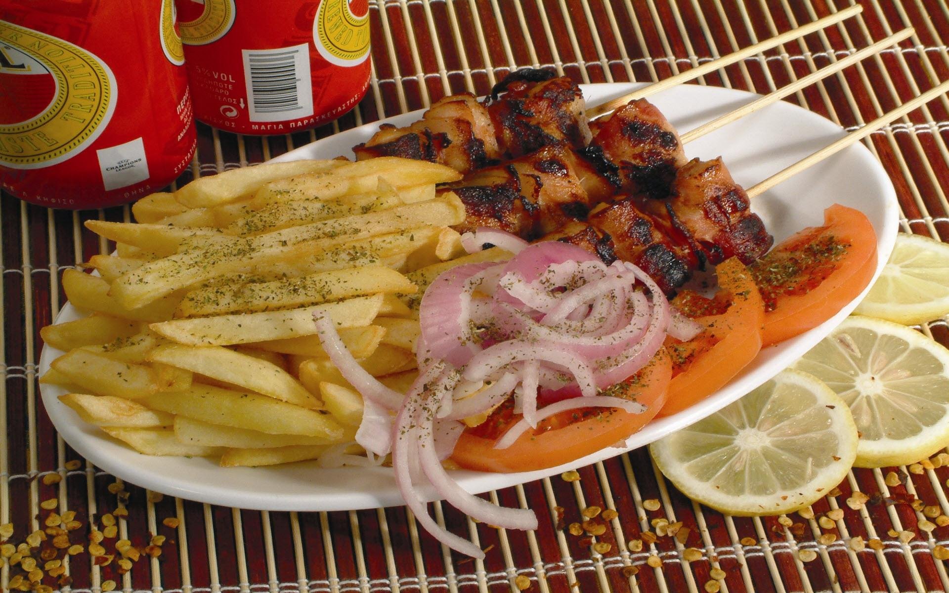 HD PC desktop wallpaper featuring a barbecue plate with grilled meat skewers, seasoned fries, sliced onions, tomatoes, and lemon wedges alongside soda cans.